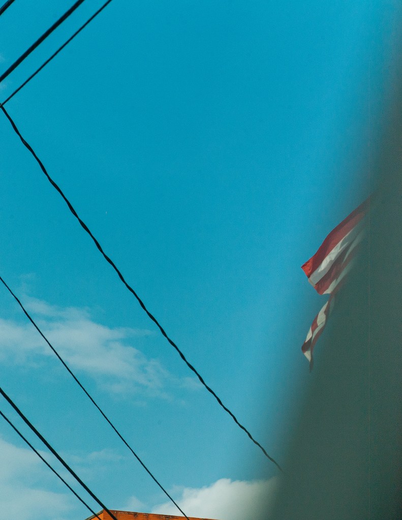 A clear blue sky with a few clouds and electrical wires crossing the image, showing part of a building and a portion of an American flag.