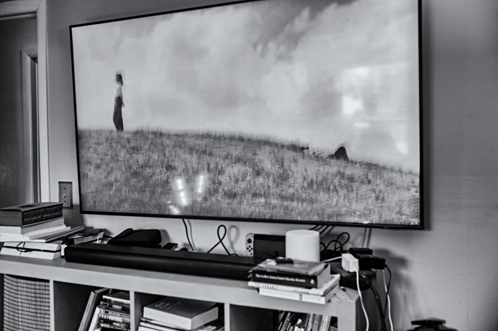 A black and white image of a person standing on a grassy hill, displayed on a television screen in a living room setting. Surrounding the TV are various books and electronic devices.