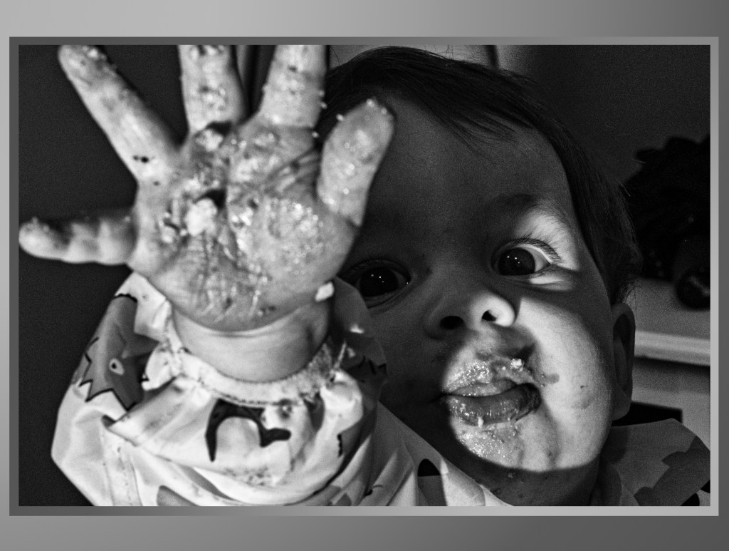 A black and white image of a young child with a playful expression, showing their hand covered in food residue, while their face is partly illuminated by light and shadow.